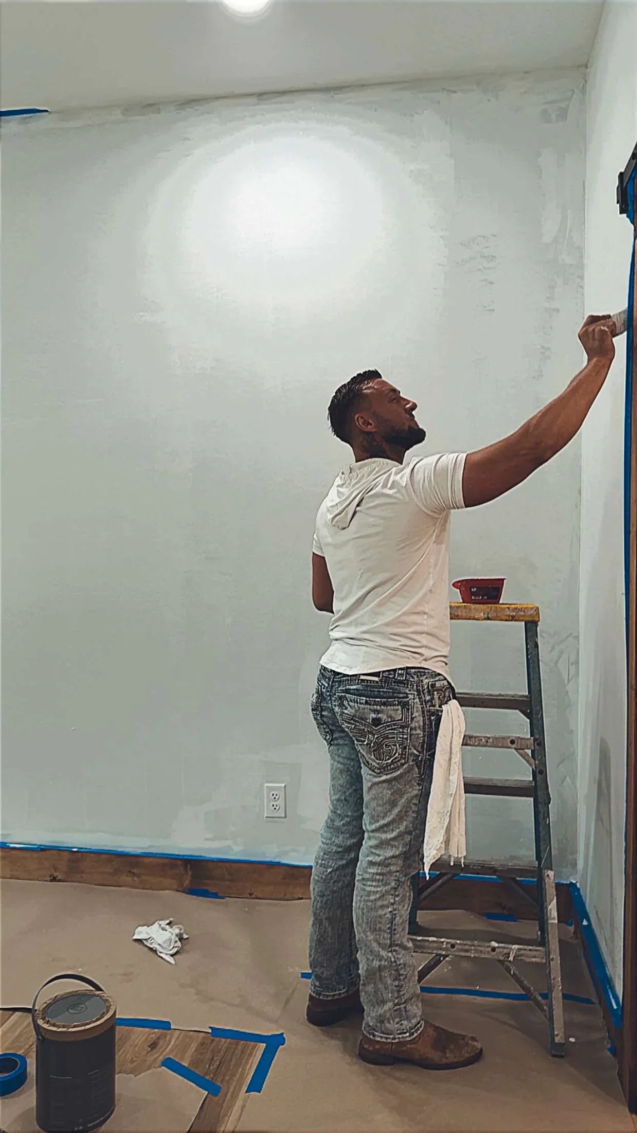 Worker on ladder applying drywall joint compound to interior wall with blue painter's tape on floor